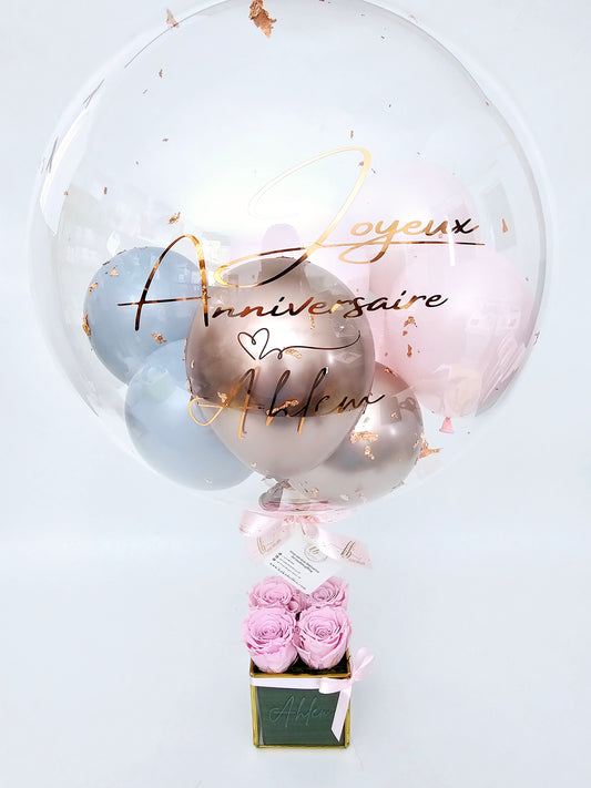 A clear balloon with printed text, floating above a square glass vase with gold frame, which contains four eternal roses. The balloon is filled with mini gold and pink balloons, and personalized with ''Joyeux anniversaite'' for a festive finish.