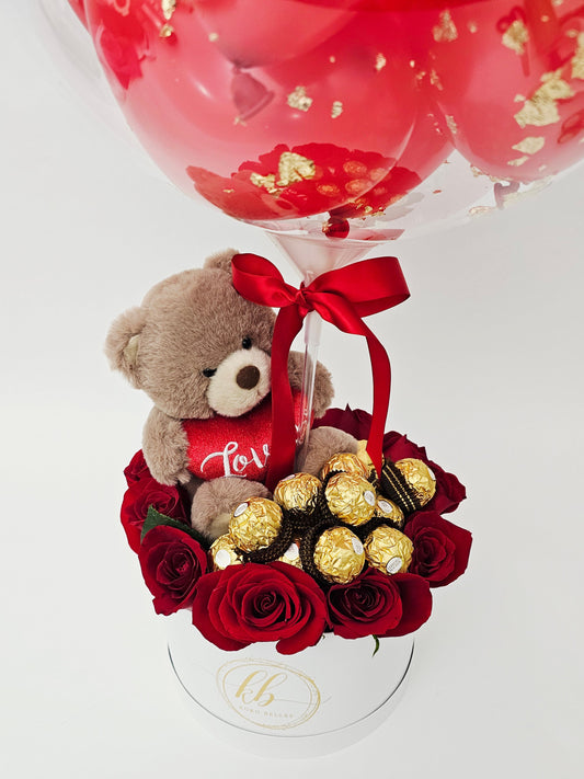 A white gift box arrangement featuring red roses, Ferrero Rocher chocolates, and a plush teddy bear holding a heart, topped with a clear bubble balloon reading "My Forever Valentine" filled with mini red balloons.