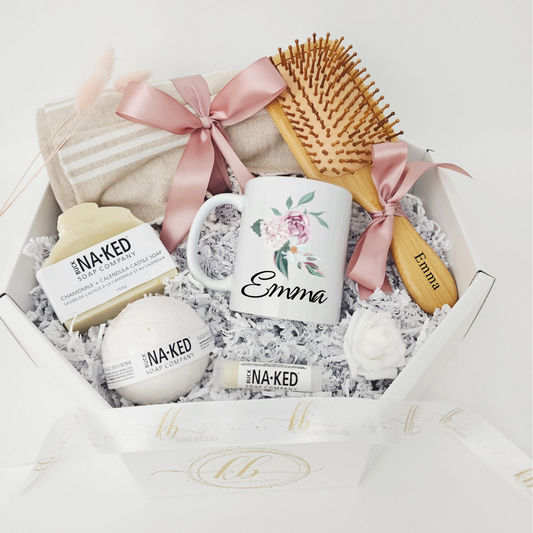 A premium Koko Belles get well soon care package featuring a personalized white ceramic mug with a delicate pink and purple floral design and the name "Emma". The white octagonal box is thoughtfully curated with a custom-engraved wooden hair brush, a luxurious linen beach towel tied with a dusty pink satin bow, and natural bath products from Buck Naked Soap Company to encourage comfort and recovery.