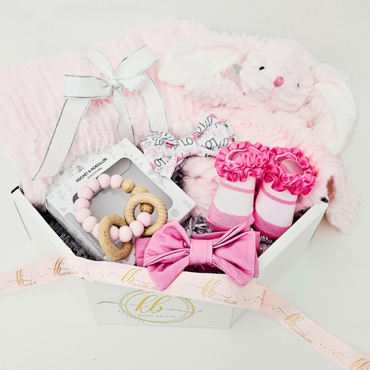 Baby gift set with pink and white items including socks, a bow, and a plush toy in a box.