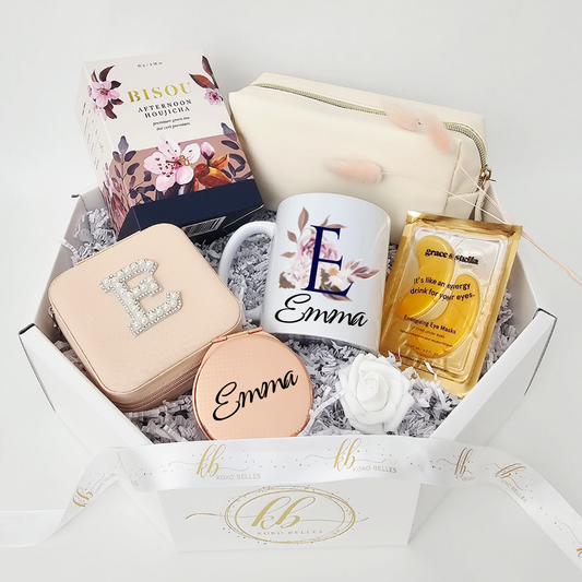A luxury bridesmaid proposal gift box presented in a white octagonal box by Koko Belles. The curated package features a personalized ceramic mug with a floral monogram 'E' and the name Emma, alongside a faux pearl initial jewelry case, rose gold compact mirror, a cream cosmetic pouch, Grace & Stella gold eye masks, and premium Bisou tea.