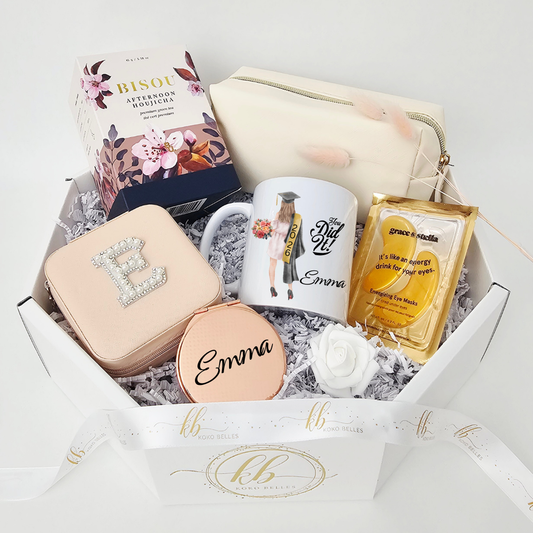 An elegant white octagonal graduation gift box featuring a custom mug with a "You Did It! 2026" graduation gown illustration personalized for Emma. The luxury care package is beautifully arranged with a pearl initial case, rose gold mirror, cosmetic bag, and gold eye masks, finished with a branded Koko Belles ribbon.