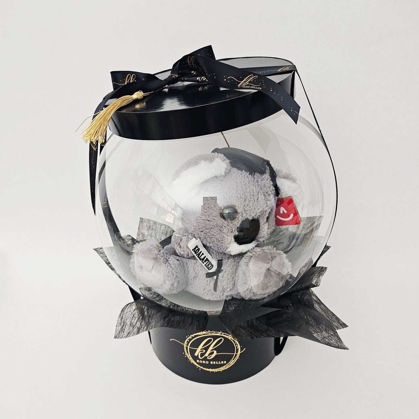 Side view of a premium air-filled transparent balloon globe enclosing a plush koala wearing a tiny graduation cap, finished with a black satin logo ribbon and a gold tassel on a black hat box base.