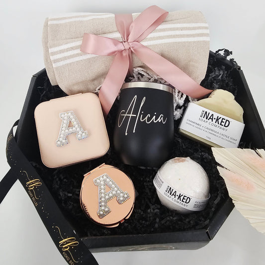 A luxury black octagonal gift box featuring a personalized matte black wine tumbler with the name Alicia in script, a blush pink initial travel jewelry case, a matching compact mirror, a beige Turkish towel, and Buck Naked soap products.