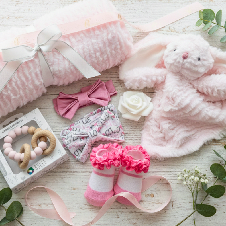Collection of baby items including a pink blanket, toy, socks, and headbands on a light wooden surface with greenery.