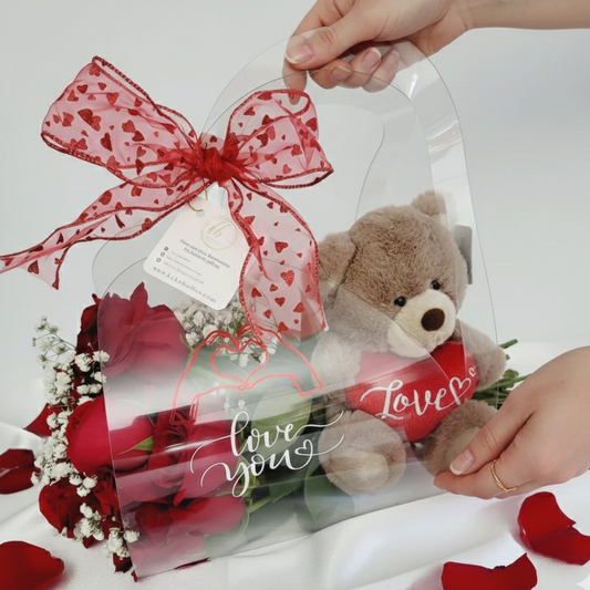A clear luxury gift tote held by hands, containing red roses and a plush brown teddy bear holding a "Love" heart.