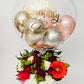Clear bubble balloon with gold 'Happy Birthday' text, metallic balloons, and a bouquet of flowers.