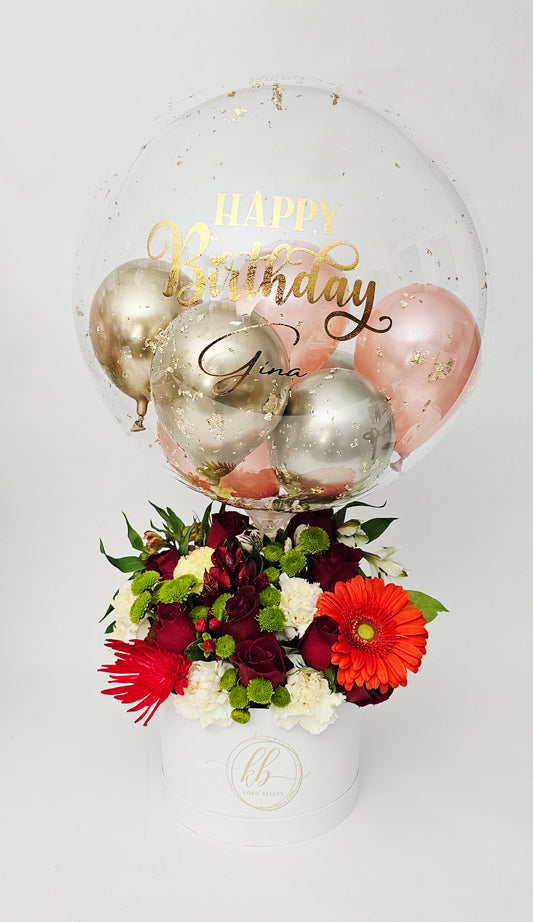 Clear bubble balloon with gold 'Happy Birthday' text, metallic balloons, and a bouquet of flowers.