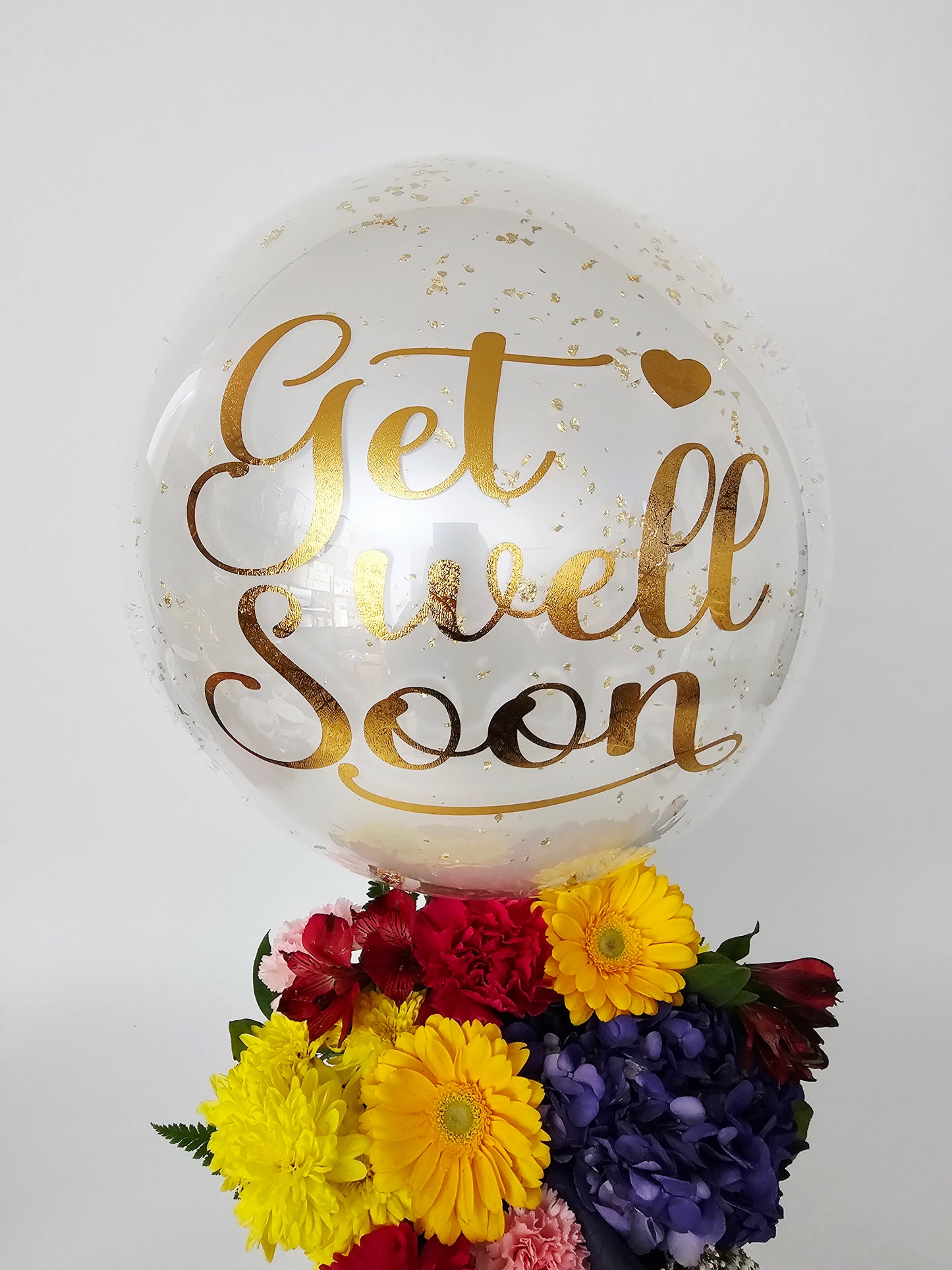 Clear balloon with gold text 'Get Well Soon' next to a bouquet of flowers on a white background
