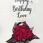 Luxury gift arrangement featuring a large, clear bubble balloon with gold script reading 'Happy Birthday Love'. The balloon is filled with gold confetti and sits above a black cylindrical hat box packed with deep red roses, a black ribbon with gold text 'KB Koko Belles', and draped black and gold-edged wrapping paper, on a white background.