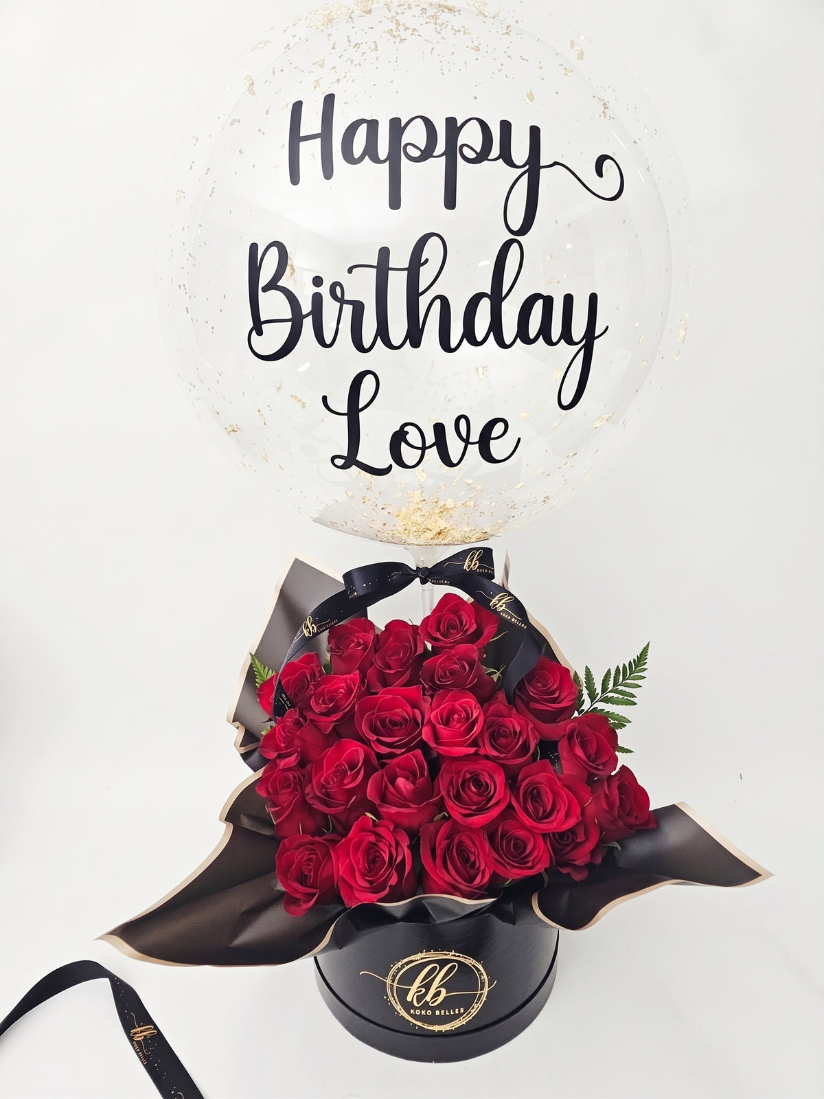 Luxury gift arrangement featuring a large, clear bubble balloon with gold script reading 'Happy Birthday Love'. The balloon is filled with gold confetti and sits above a black cylindrical hat box packed with deep red roses, a black ribbon with gold text 'KB Koko Belles', and draped black and gold-edged wrapping paper, on a white background.