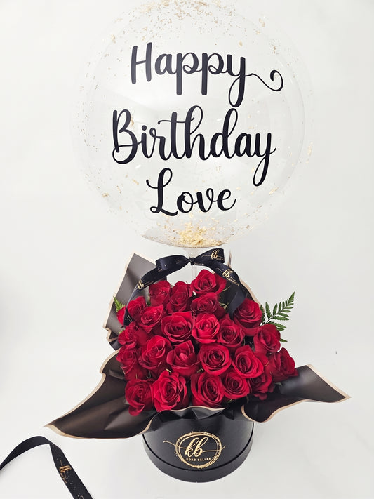 Luxury gift arrangement featuring a large, clear bubble balloon with gold script reading 'Happy Birthday Love'. The balloon is filled with gold confetti and sits above a black cylindrical hat box packed with deep red roses, a black ribbon with gold text 'KB Koko Belles', and draped black and gold-edged wrapping paper, on a white background.