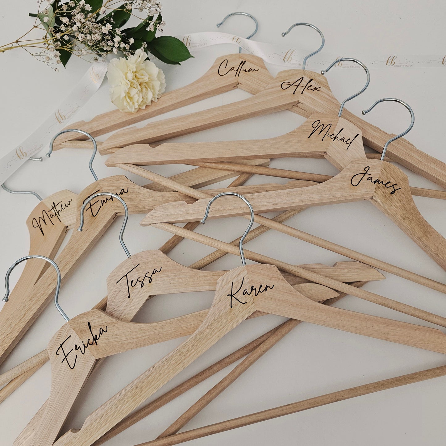 Wooden hangers with names engraved on them, arranged on a white surface with flowers.