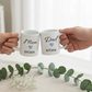 Two white mugs with 'Mom EST. 2026' and 'Dad EST. 2026' text held by hands on a light surface with greenery.