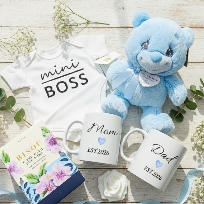Baby-themed items including a 'mini BOSS' onesie, blue teddy bear, 'Mom' and 'Dad' mugs, and a box of tea on a wooden surface.