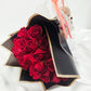 Un élégant bouquet de roses rouges enveloppé dans un papier noir et or, présenté dans un sac cadeau transparent personnalisé avec un petit ourson en peluche.