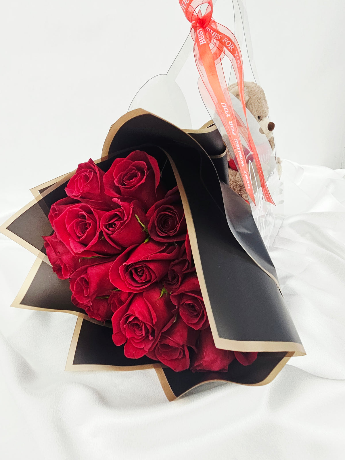 Un élégant bouquet de roses rouges enveloppé dans un papier noir et or, présenté dans un sac cadeau transparent personnalisé avec un petit ourson en peluche.