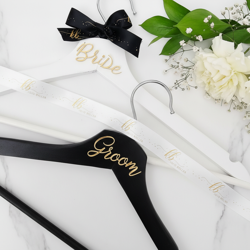 Black hangers with 'Bride' and 'Groom' text on a white background with flowers.