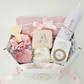 A luxury white octagonal gift box filled with blush pink and rose gold personalized items, including a white skinny tumbler customized with the name Anna, a pink jewelry case, a hair scrunchie, and gold hair accessories on white shredded paper.