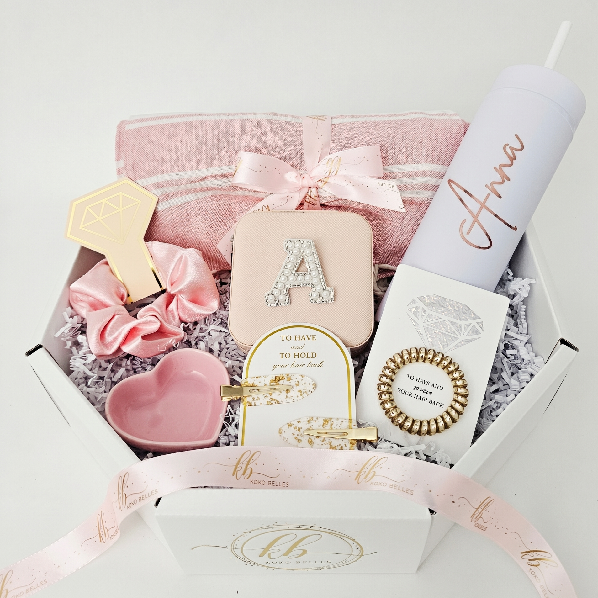A luxury white octagonal gift box filled with blush pink and rose gold personalized items, including a white skinny tumbler customized with the name Anna, a pink jewelry case, a hair scrunchie, and gold hair accessories on white shredded paper.