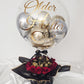 Personalized clear bubble balloon with gold script reading 'Older Hotter Better', filled with silver and gold mini balloons, set atop a black hat box filled with red roses and Ferrero Rocher chocolates.