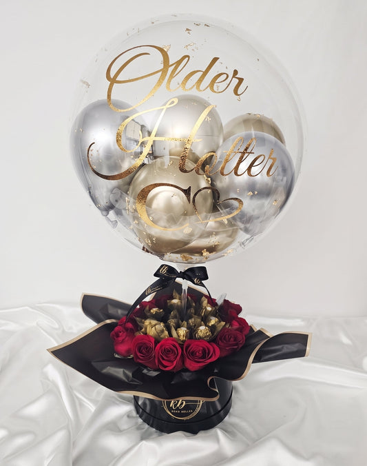 Personalized clear bubble balloon with gold script reading 'Older Hotter Better', filled with silver and gold mini balloons, set atop a black hat box filled with red roses and Ferrero Rocher chocolates.