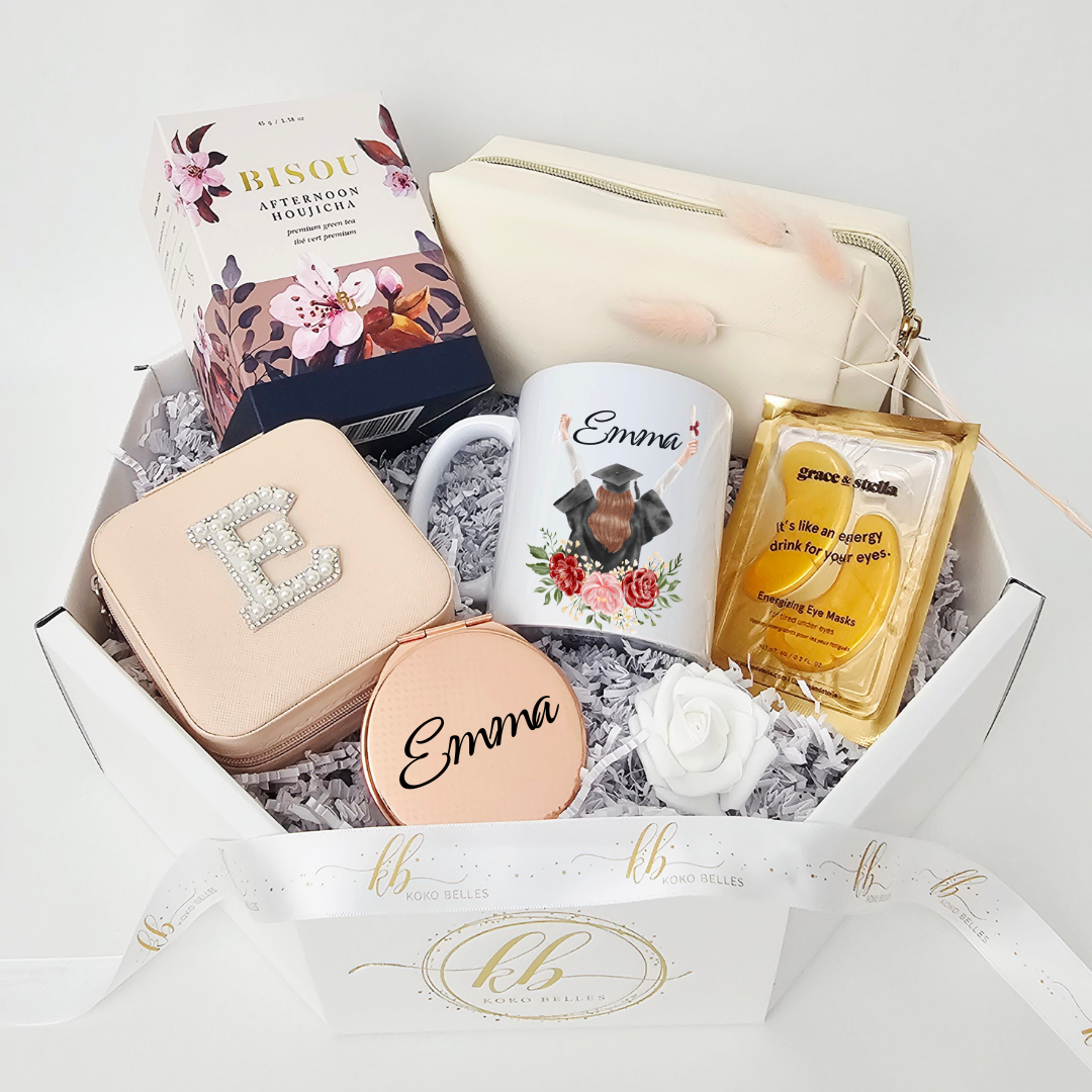 A luxury white octagonal graduation care package by Koko Belles featuring a personalized mug with a floral graduation girl illustration and the name Emma. The gift box includes a pearl initial jewelry case, rose gold compact mirror, premium Bisou Houjicha tea, a cream cosmetic pouch, and Grace & Stella eye masks over white crinkle paper.