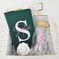 A straight overhead view of a premium open gift box by Koko Belles, showcasing a matching personalized stationery and drinkware set. The care package includes a custom monogrammed emerald green notebook, a clear glass can tumbler with a natural bamboo lid and glass straw, a white rose decoration, and elegant gold heart tissue paper lining.