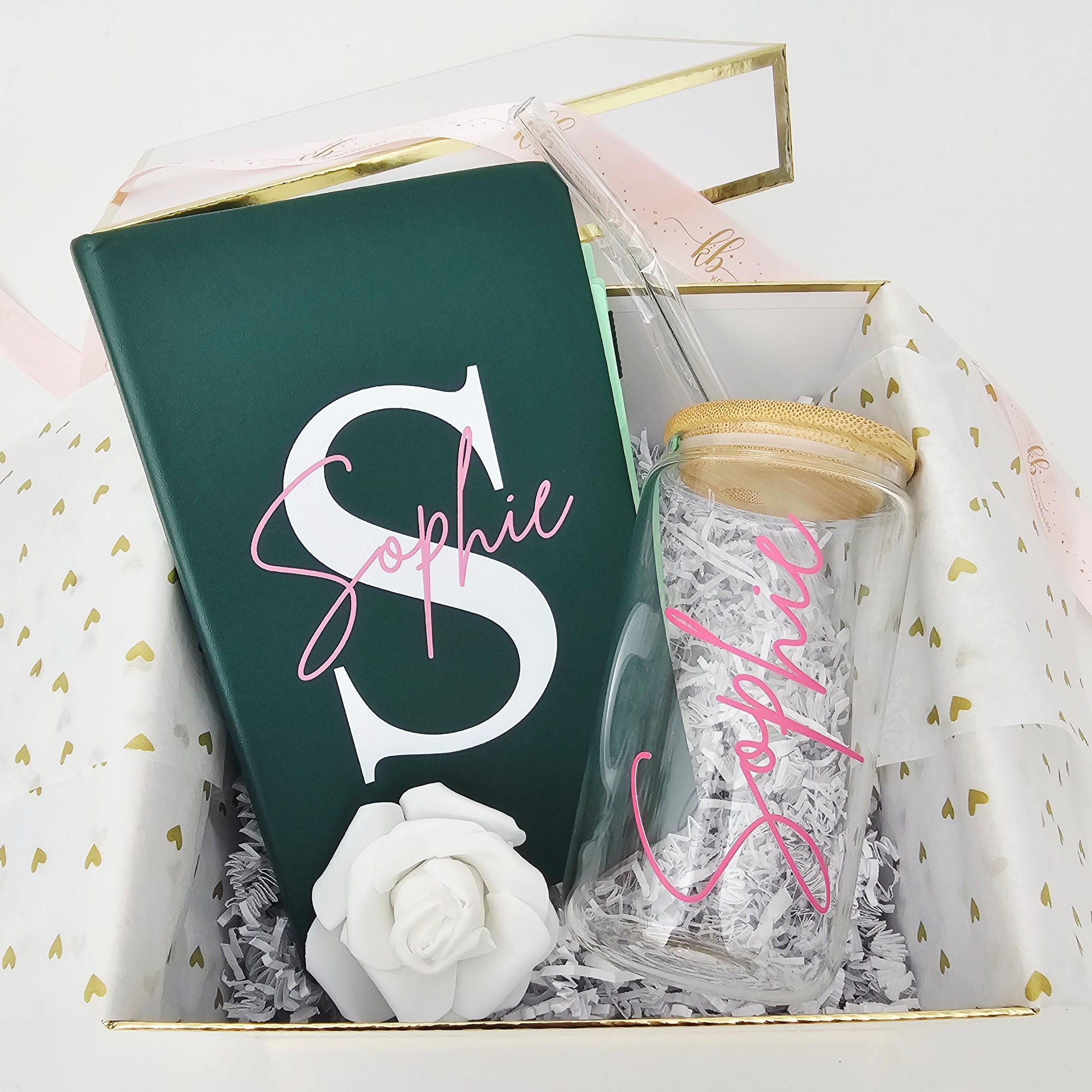 A straight overhead view of a premium open gift box by Koko Belles, showcasing a matching personalized stationery and drinkware set. The care package includes a custom monogrammed emerald green notebook, a clear glass can tumbler with a natural bamboo lid and glass straw, a white rose decoration, and elegant gold heart tissue paper lining.