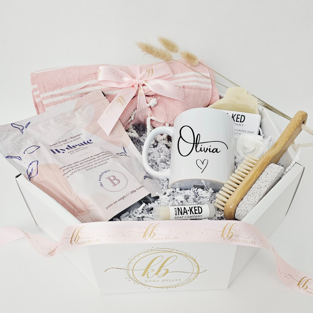 An angled view of a premium custom wellness care package in a white octagonal presentation box. The beautifully curated self-care set showcases a personalized name mug alongside natural bath products, an eco-friendly wooden nail and pumice brush, a pink cotton beach towel, and a branded Koko Belles satin ribbon.
