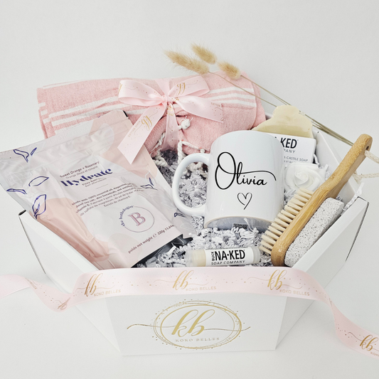 An angled view of a premium custom wellness care package in a white octagonal presentation box. The beautifully curated self-care set showcases a personalized name mug alongside natural bath products, an eco-friendly wooden nail and pumice brush, a pink cotton beach towel, and a branded Koko Belles satin ribbon.