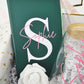 A detailed view focusing on a premium emerald green faux-leather notebook customized with a bold white monogram letter "S" and the name "Sophie" in elegant pink script. The journal is nestled inside a luxury Koko Belles gift box alongside a white rose accent and a clear glass straw.