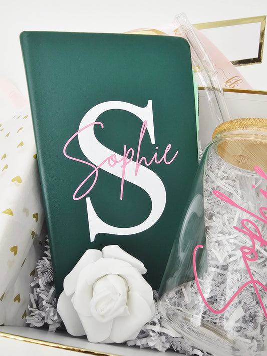 A detailed view focusing on a premium emerald green faux-leather notebook customized with a bold white monogram letter "S" and the name "Sophie" in elegant pink script. The journal is nestled inside a luxury Koko Belles gift box alongside a white rose accent and a clear glass straw.