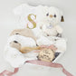 Luxury personalized baby girl gift basket featuring a gold glitter initial onesie, engraved wooden hairbrush, and birth announcement plaque in a white gift box.