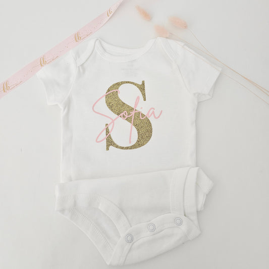 Personalized baby girl onesie with large sparkly gold glitter initial S and pink script name Sofia.