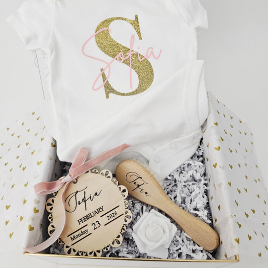 The ultimate KokoBelles gift set in a gold-trimmed white box featuring a personalized onesie, engraved hairbrush, and birth announcement plaque.