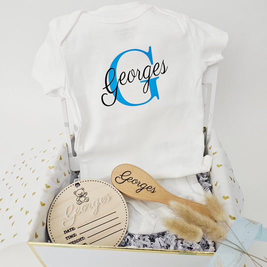 A luxury KokoBelles baby boy gift set in a gold-trimmed box featuring a blue initial onesie, personalized wooden brush, and engraved birth announcement plaque.