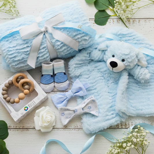 Baby gift set with blue teddy bear, booties, and other items on a wooden surface.
