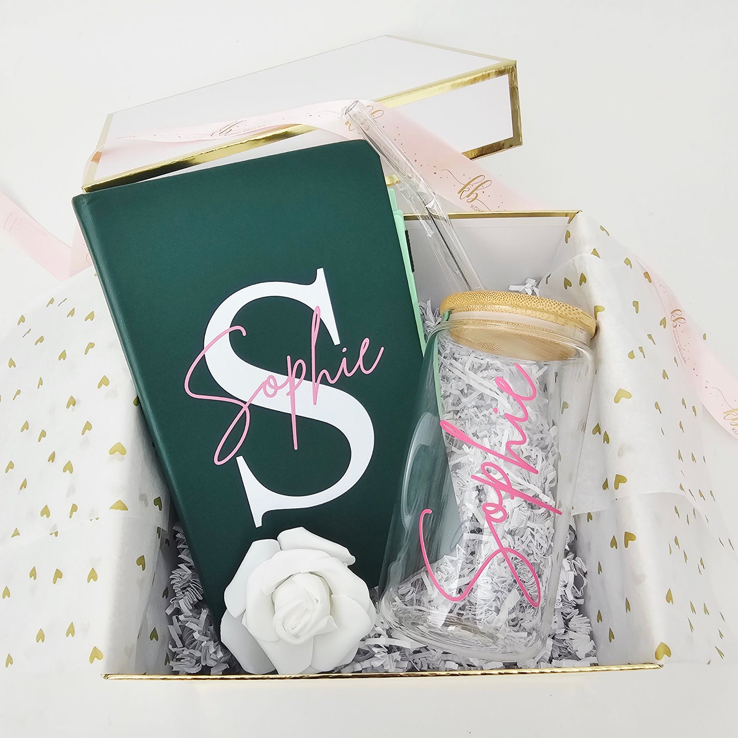 An angled, full view of an open Koko Belles luxury gift box lined with gold trim and white tissue paper patterned with gold hearts. The curated set features a personalized emerald green journal and a custom glass can tumbler with a bamboo lid, both featuring the name "Sophie" in pink, complete with a signature Koko Belles branded satin ribbon resting on the lid.