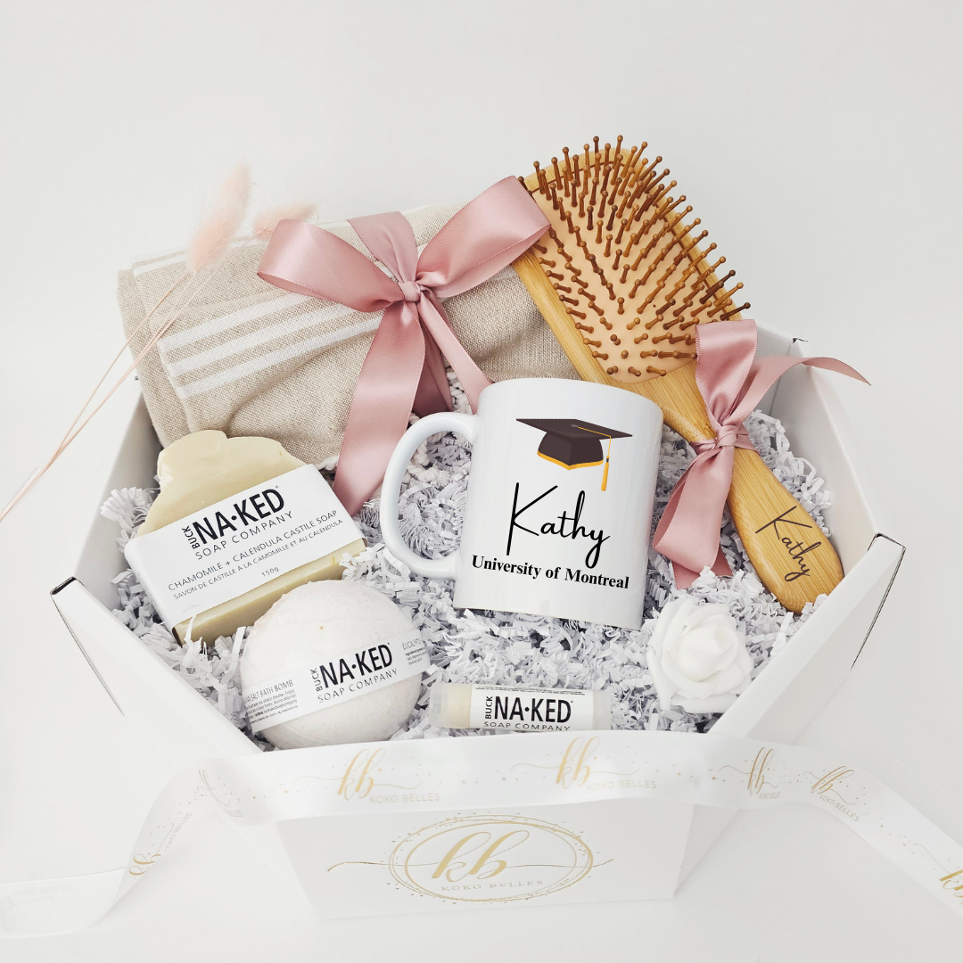 A luxury graduation self-care gift basket featuring a custom University of Montreal mug with the name Kathy, an engraved wooden hairbrush, a linen hand towel with a pink satin bow, and Buck Naked soap products in a white octagonal box.