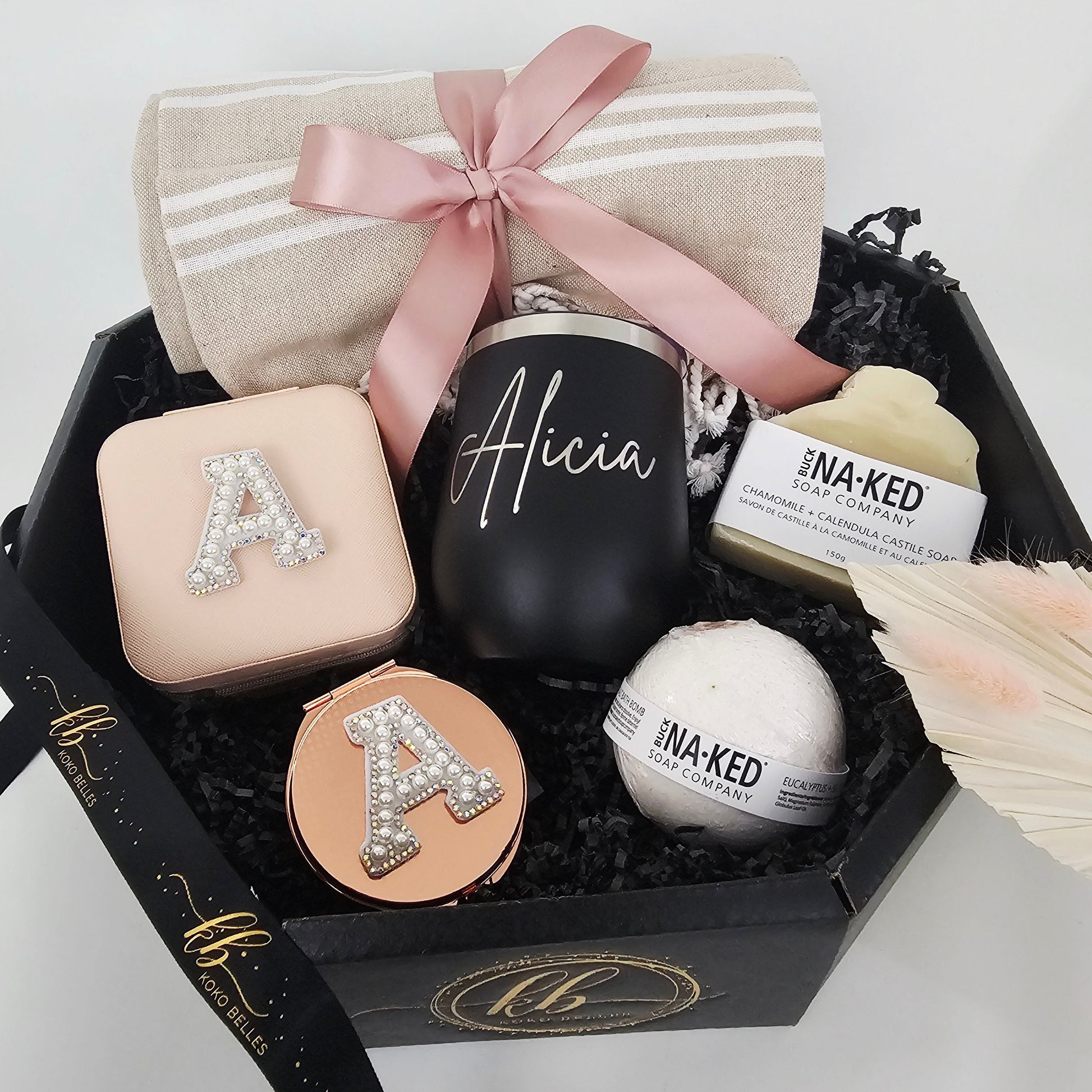 An elegant custom gift box with black shredded paper fill, containing a personalized initials jewelry box, compact mirror, a black tumbler, natural bar soap, and a bath bomb.