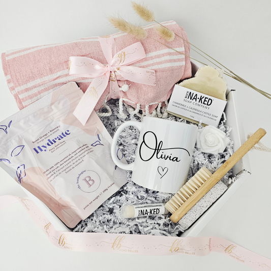 A luxury white octagonal spa care package by Koko Belles featuring a personalized ceramic mug with the name Olivia and a small heart. The custom gift basket includes a blush pink beach towel tied with a ribbon, Buck Naked Soap Company castile soap and lip balm, The Bathologist "Hydrate" bath soak, a wooden foot brush, and a white rose over crinkle paper.