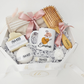 An overhead view of a Koko Belles self-care gift box designed for relaxation, highlighting a custom white mug with minimalist boho shapes in mustard and terracotta with fine line art flowers and the name "Emma". The curated box contains premium wellness essentials, including a custom-engraved bamboo hair brush, a soft linen beach towel, and artisanal Buck Naked Soap Company skincare products on clean white crinkle paper.