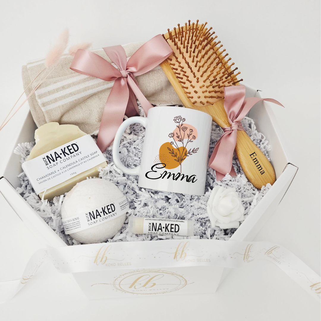 An overhead view of a Koko Belles self-care gift box designed for relaxation, highlighting a custom white mug with minimalist boho shapes in mustard and terracotta with fine line art flowers and the name "Emma". The curated box contains premium wellness essentials, including a custom-engraved bamboo hair brush, a soft linen beach towel, and artisanal Buck Naked Soap Company skincare products on clean white crinkle paper.