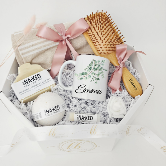 A high-end custom gift for her by Koko Belles, beautifully arranged in an elegant white octagonal box. The luxury set features a personalized mug with a soft watercolor eucalyptus leaf design and the name "Emma", alongside a laser-engraved wooden paddle brush, a premium beach towel wrapped in a dusty pink satin ribbon, and natural Buck Naked Soap Company bath treats.