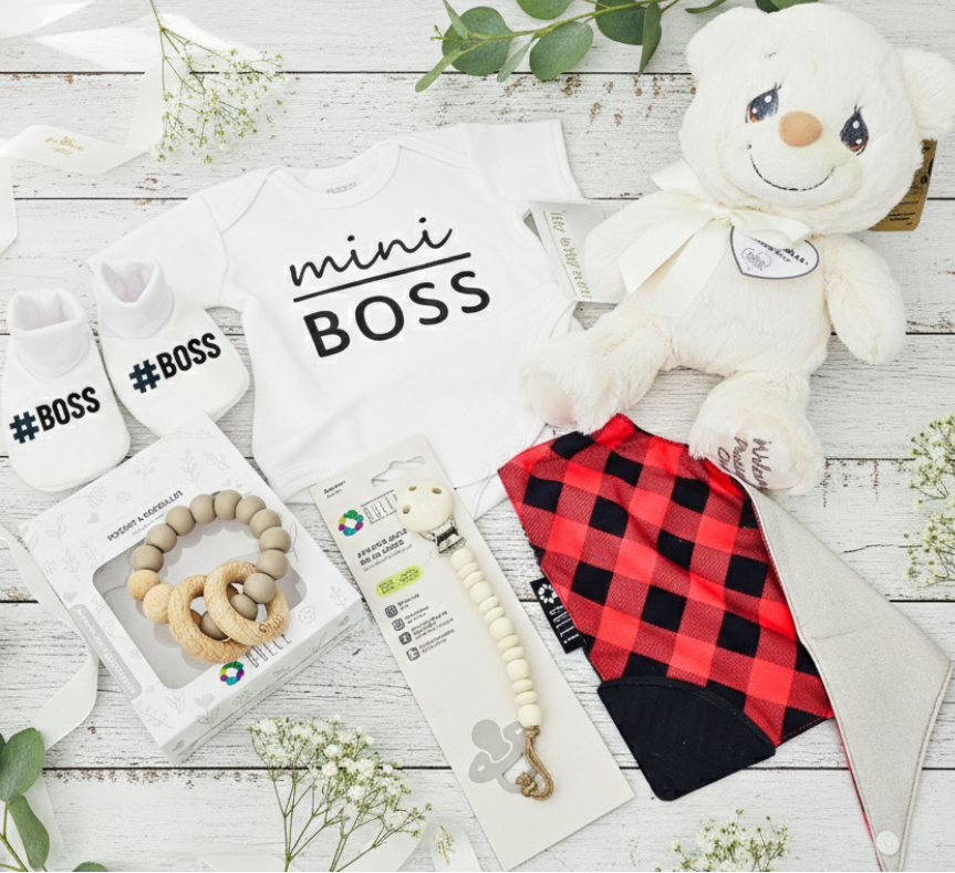 Baby items including a teddy bear, baby clothes, and accessories on a wooden surface with greenery.