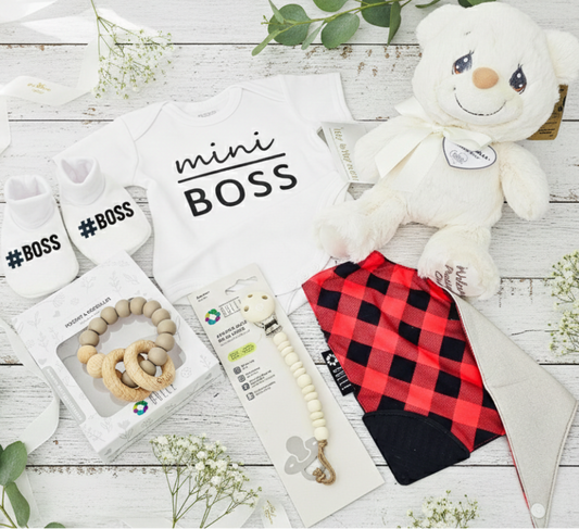 Baby items including a teddy bear, baby clothes, and accessories on a wooden surface with greenery.