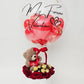 A luxury Valentine's Day gift box with red roses, gold chocolates, a teddy bear, and a custom script bubble balloon.