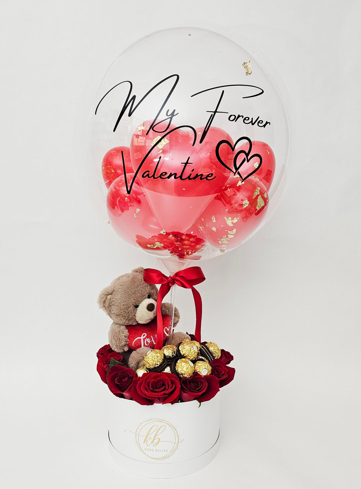 A luxury Valentine's Day gift box with red roses, gold chocolates, a teddy bear, and a custom script bubble balloon.