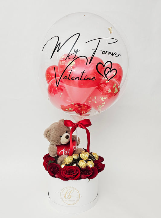 A luxury Valentine's Day gift box with red roses, gold chocolates, a teddy bear, and a custom script bubble balloon.